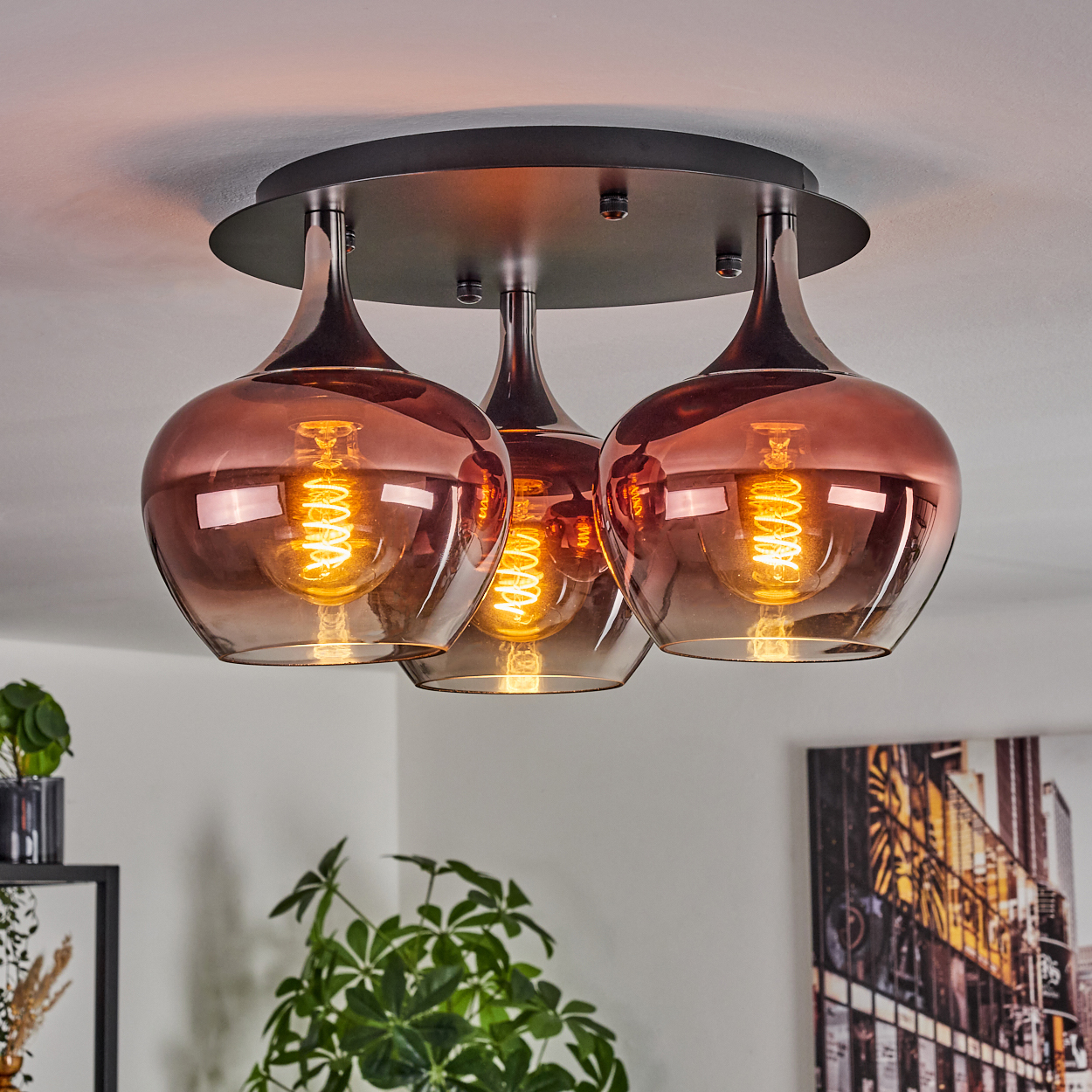 Eine moderne Deckenlampe mit drei rosafarbenen Glasschirmen. Warme Glühbirnen beleuchten die Schirme, mit Pflanzen im Vordergrund.