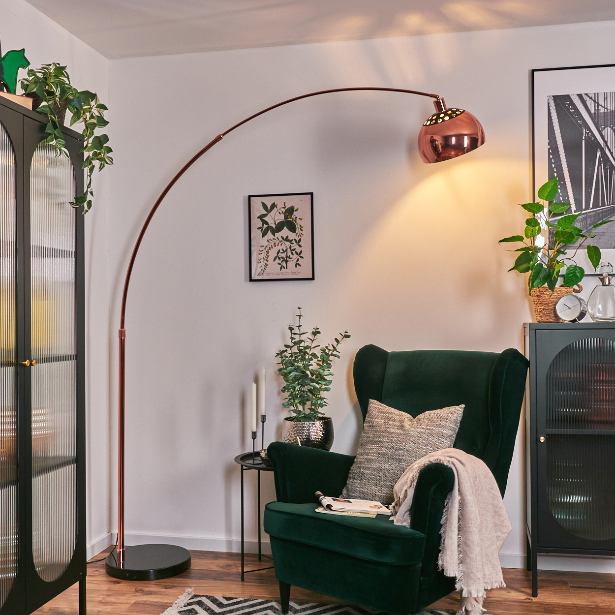 Ein grüner Sessel mit Kissen und Decke steht unter einer kupferfarbenen Stehlampe. Pflanzen und ein schwarzer Schrank sind im Hintergrund.
