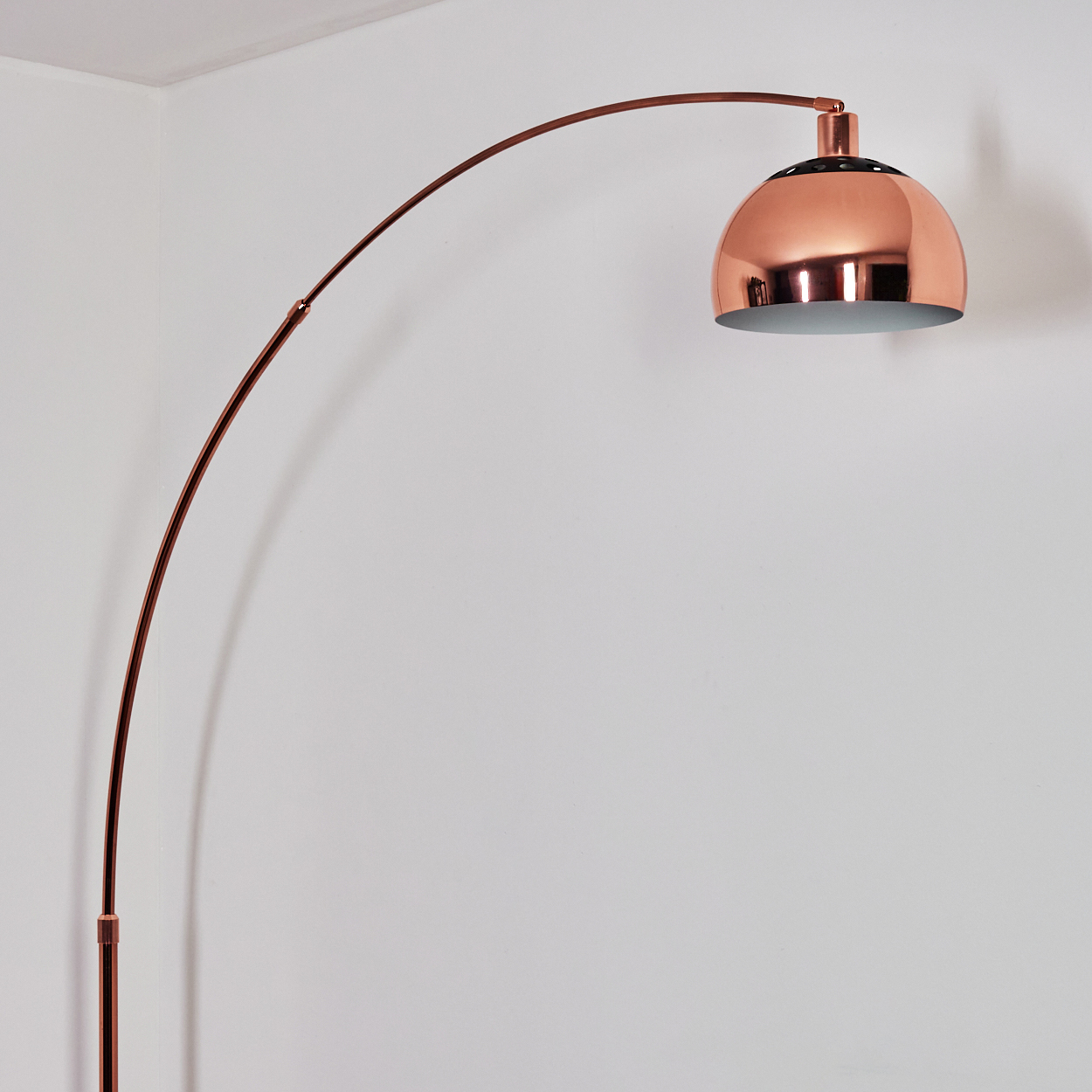Eine kupferfarbene Bogenstehlampe steht vor einer weißen Wand. Die Lampe hat einen gebogenen Arm und einen kuppelförmigen Schirm.