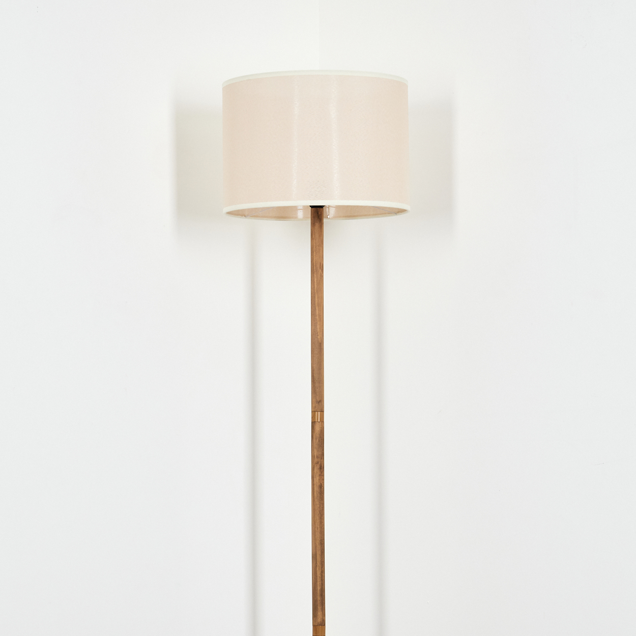 Eine Stehlampe steht vor einer weißen Wand. Die Lampe hat einen beigen Lampenschirm und einen Holzpfahl.