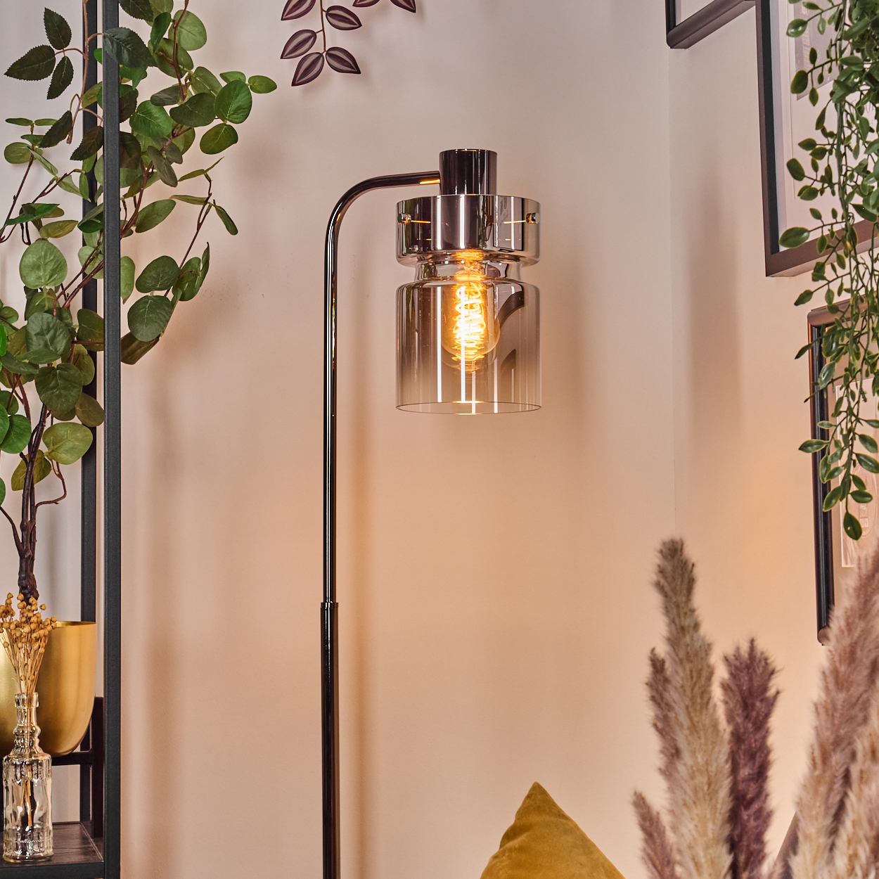 Eine moderne Stehlampe mit leuchtender Glühbirne. Grüne Pflanzen und Dekoratives umgeben sie, vor einer cremefarbenen Wand.