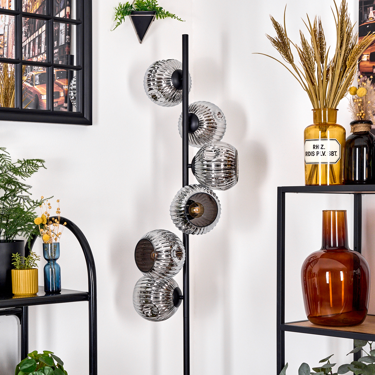 Eine moderne Stehlampe mit Glaskugeln vor einer weißen Wand. Regale mit Vasen und Dekoration sind sichtbar. Ein gerahmtes Bild hängt.