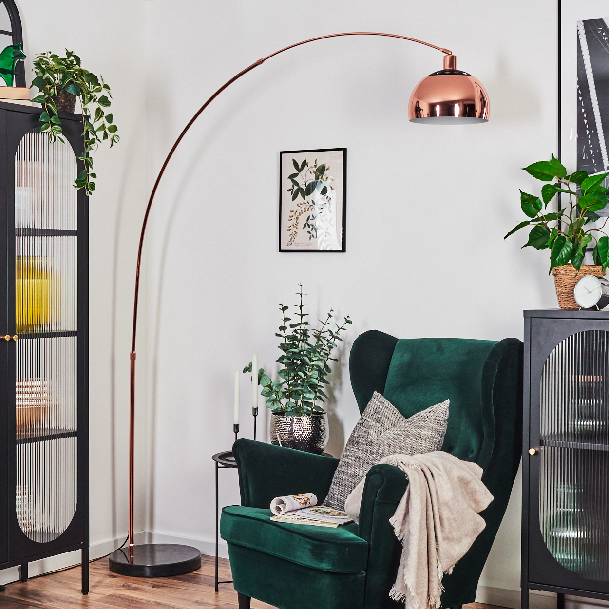 Ein Zimmer mit grünem Sessel, Kupferlampe, schwarzen Schränken und Pflanzen. Ein Bild hängt an der Wand, und eine Decke liegt auf dem Stuhl.