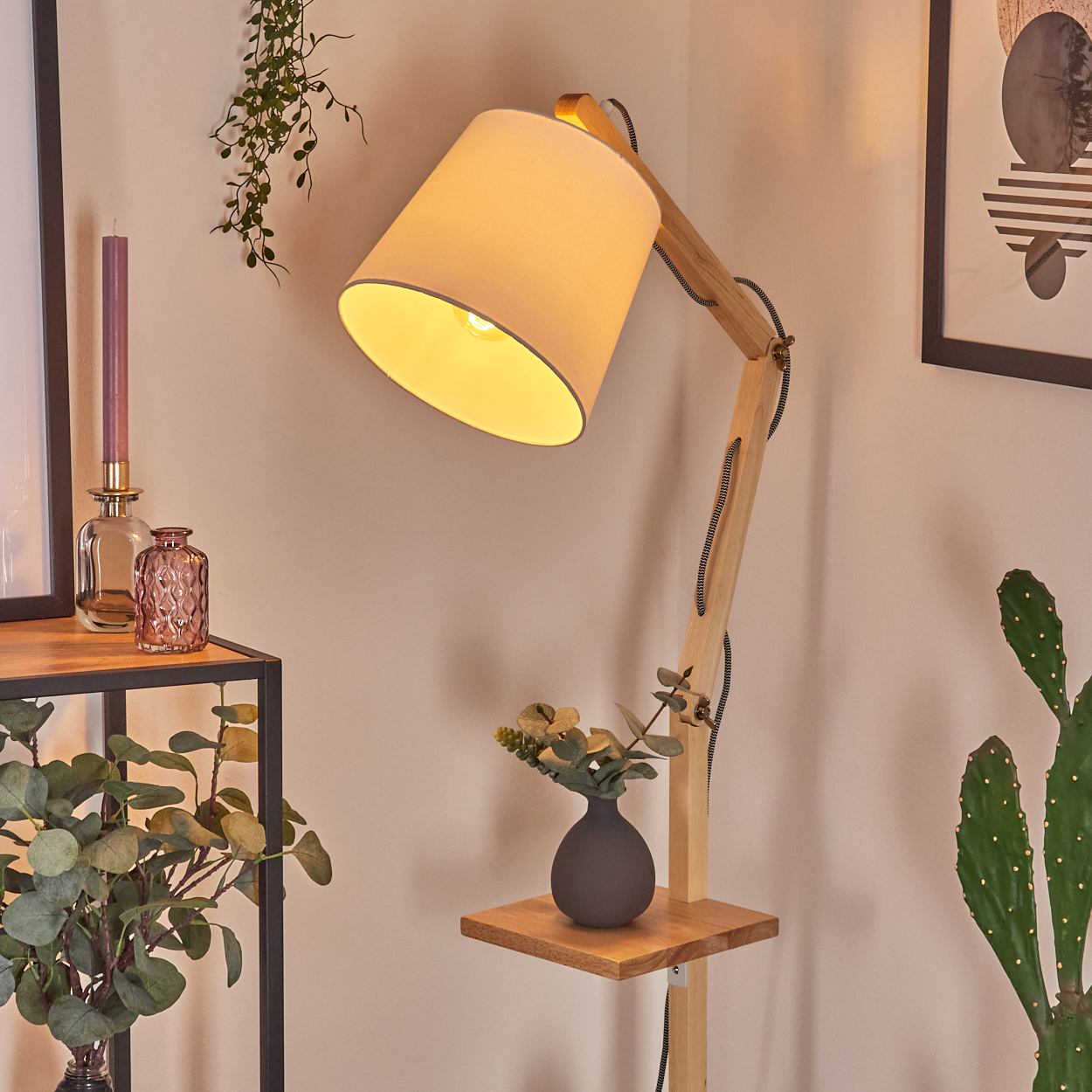 Eine Holz-Stehlampe mit beigem Schirm steht in einem Raum. Ein Regal hält eine Vase mit Blumen. Rechts ist ein Kaktus.