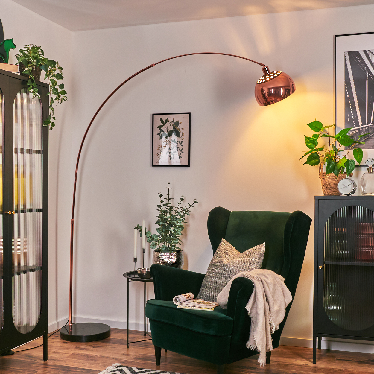 Ein grüner Sessel mit Decke und Kissen, in der Nähe einer Stehlampe. Links ein Schrank mit Pflanzen. Ein Bild an der Wand.