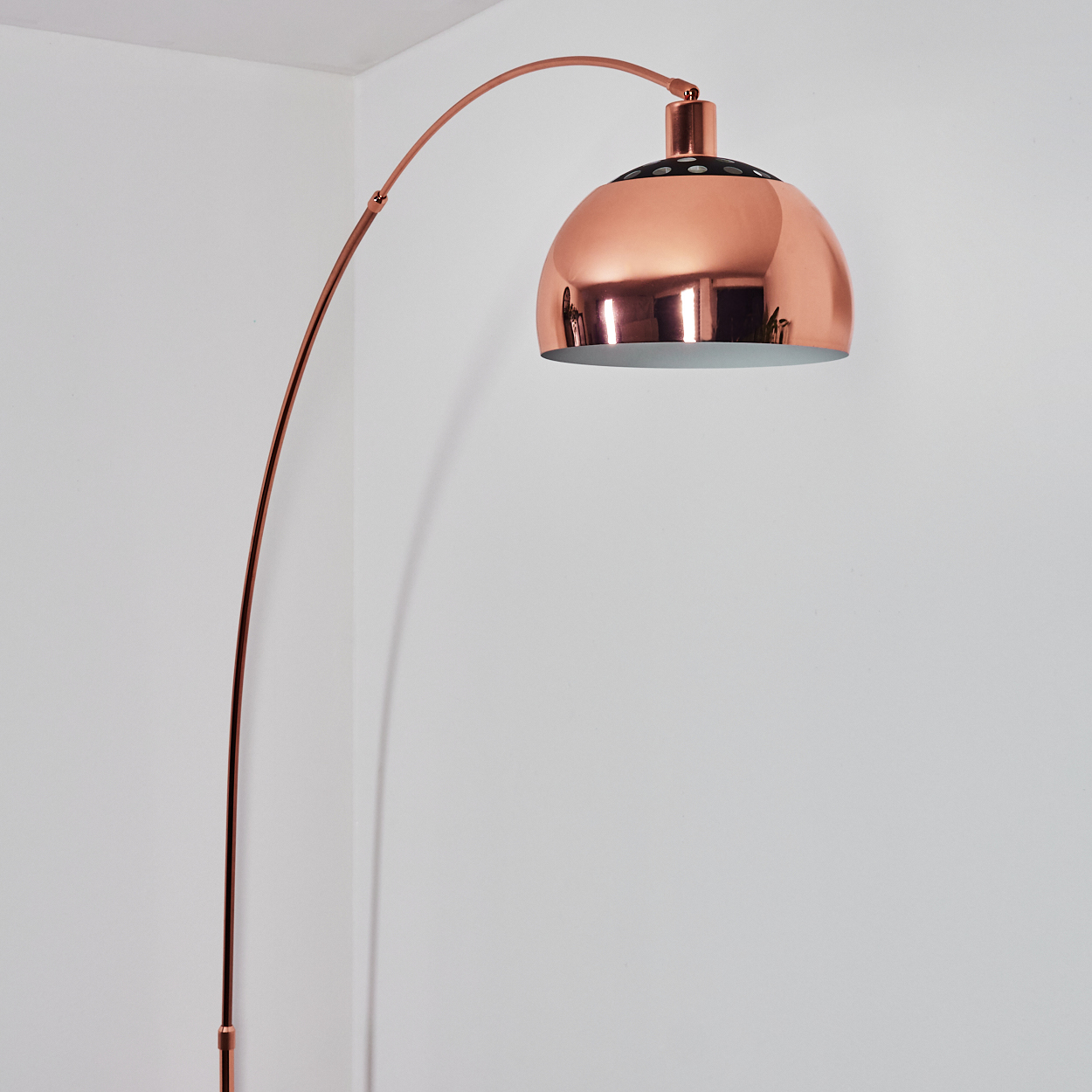 Eine kupferfarbene Bogenstehlampe steht vor einer weißen Wand. Die Lampe hat einen großen, runden Schirm und einen gebogenen Stiel.