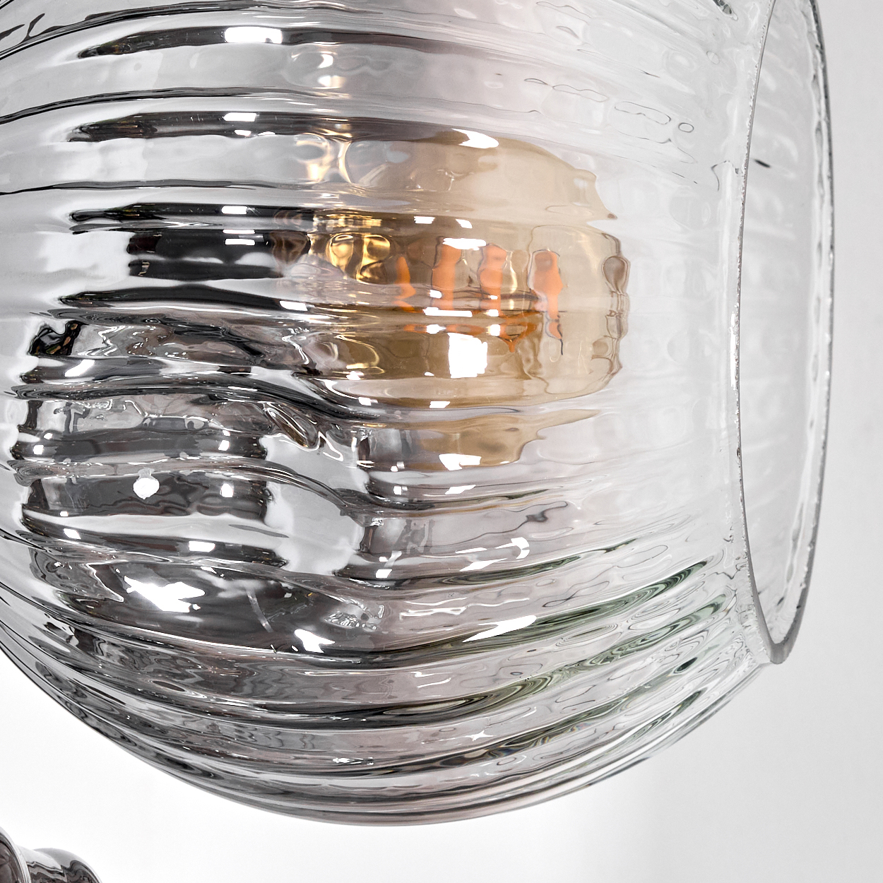 Nahaufnahme eines Lampenschirms aus klarem Glas mit gewellter Textur, der eine warme Glühbirne im Inneren zeigt, vor weißem Hintergrund.