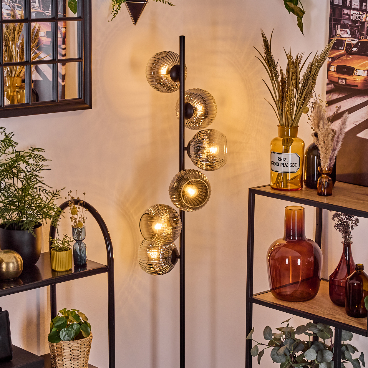 Ein modernes Interieur mit einer Stehlampe mit kugelförmigen Glasschirmen, Regalen mit Pflanzen und Vasen sowie einem Bild.