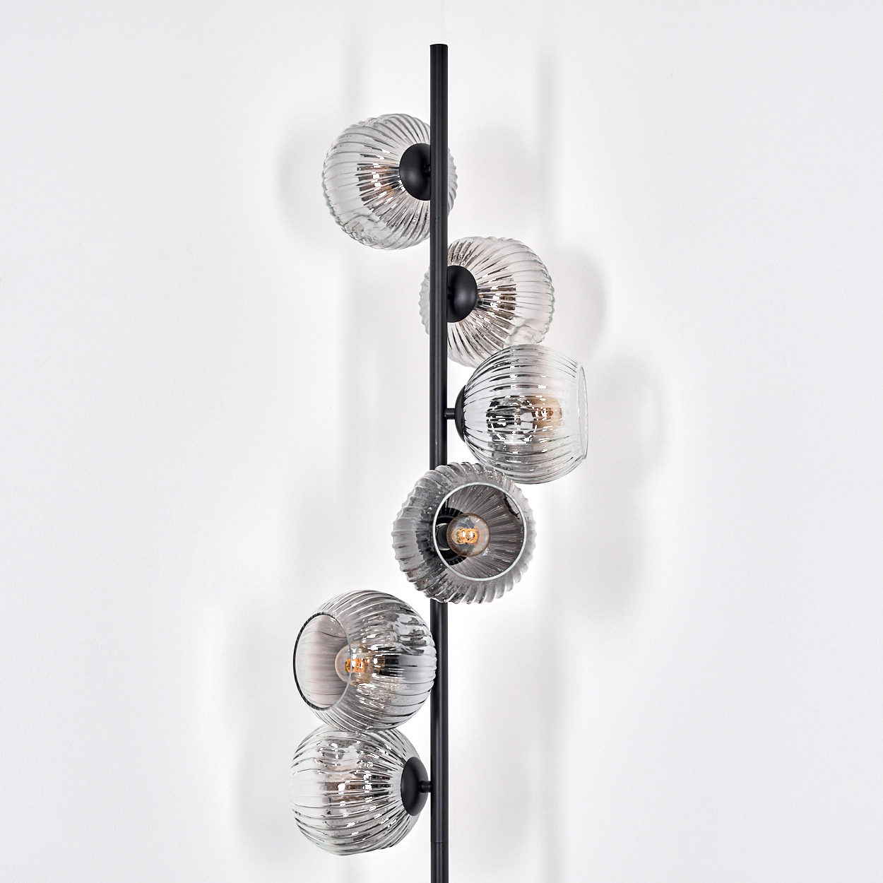 Eine moderne schwarze Stehlampe mit sechs Glaskugelschirmen. Jedes hat eine gerippte Textur und enthält eine Glühbirne. Sie steht vor einer weißen Wand.