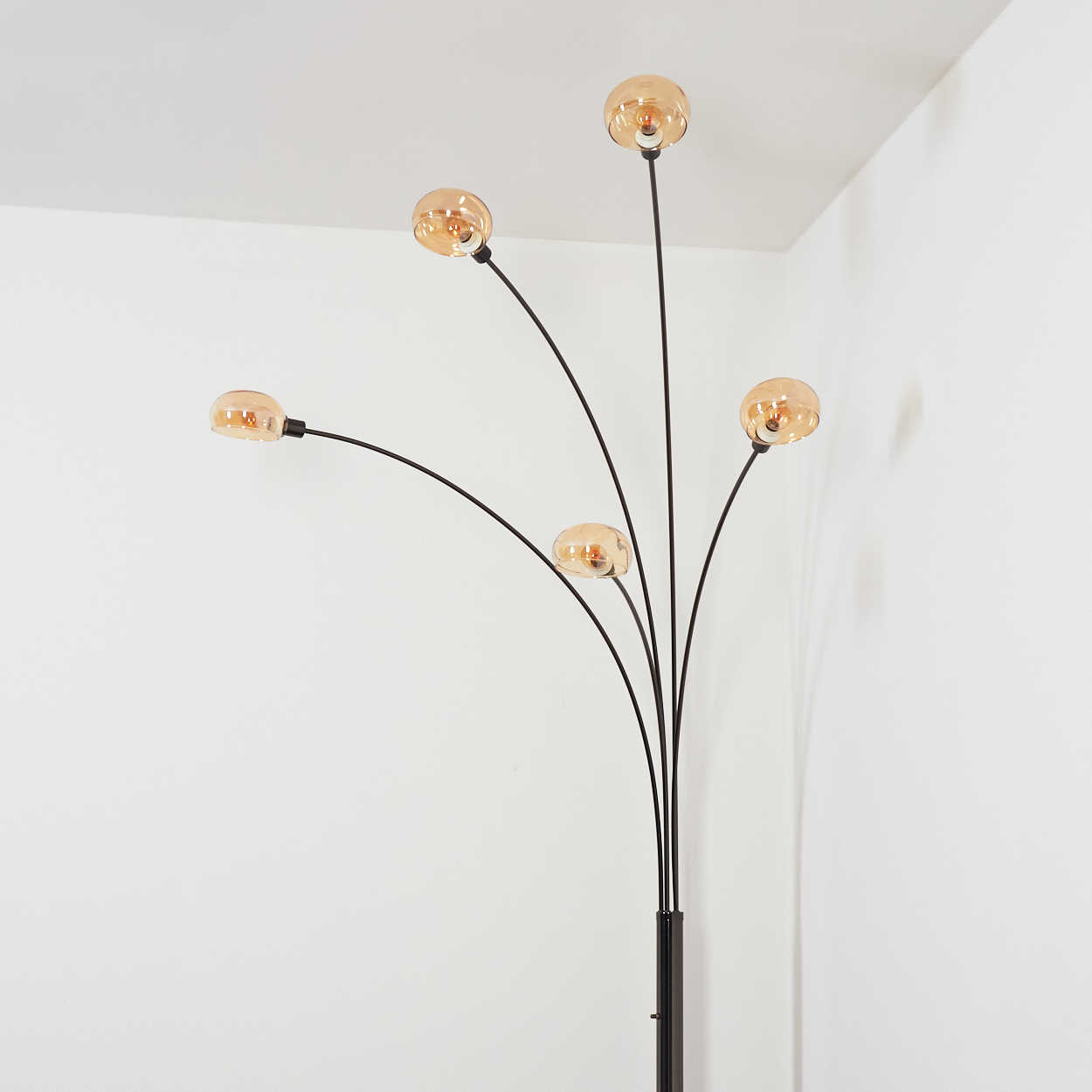 Eine moderne Stehlampe mit fünf bernsteinfarbenen Kugeln. Die Lampe hat ein schwarzes, gewölbtes Gestell vor einer weißen Wand und Ecke.