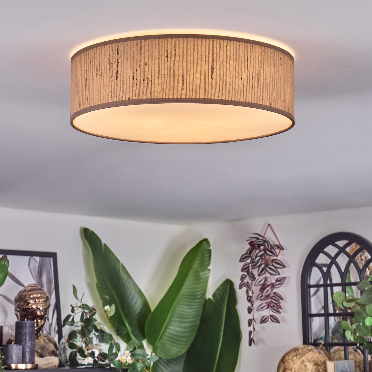 Eine Deckenlampe mit beigem Schirm ist zentriert. Große grüne Blätter und Dekoration befinden sich darunter. Der Hintergrund ist eine weiße Wand.