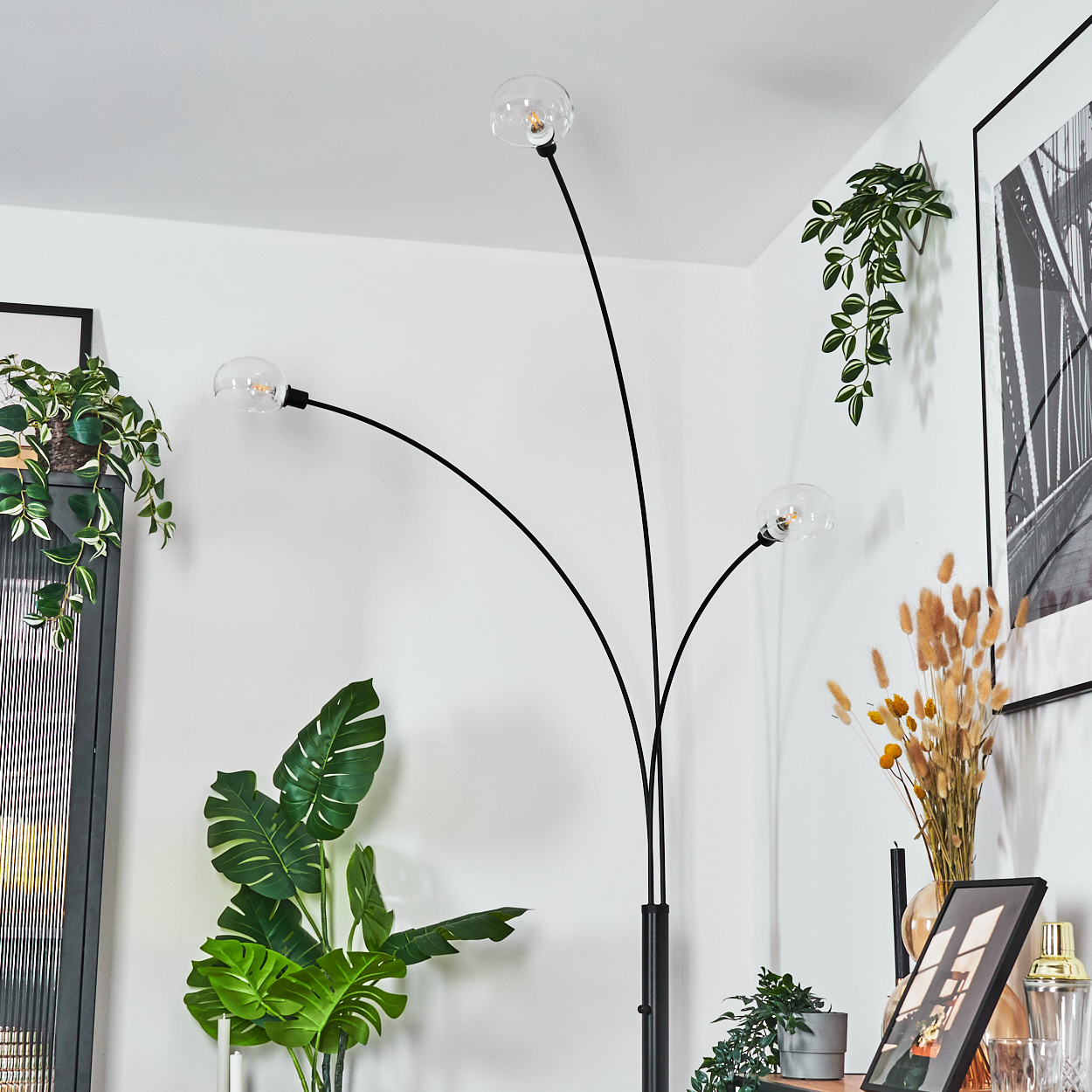 Eine moderne Stehlampe mit drei Glaskugelleuchten. Pflanzen und gerahmte Kunstwerke umgeben sie. Der Hintergrund ist eine weiße Wand.