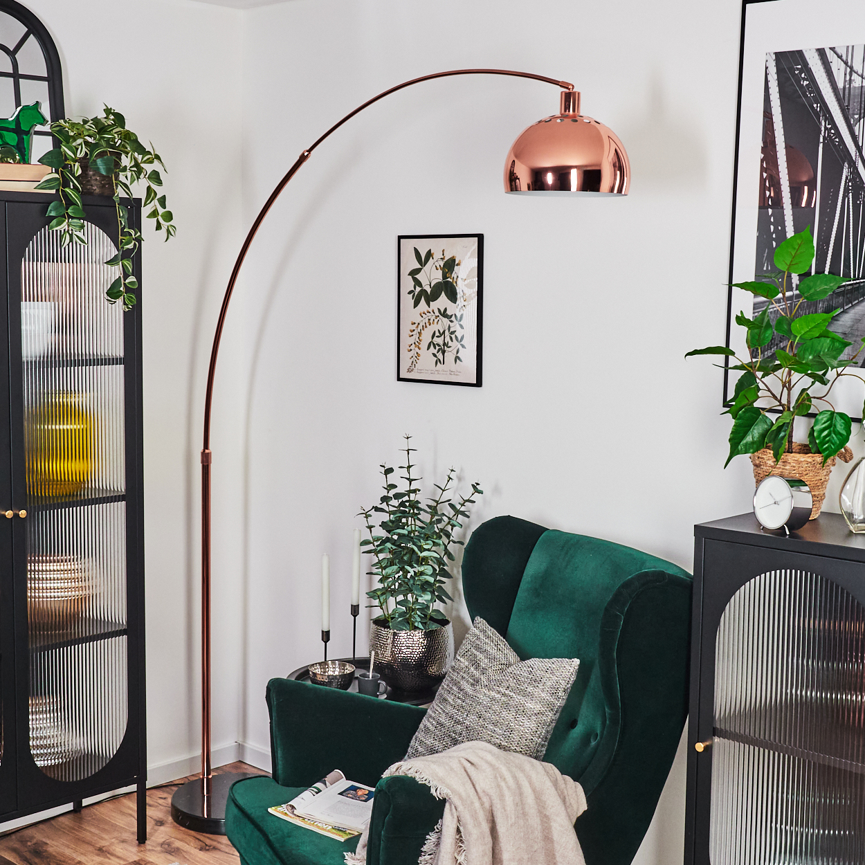 Ein Raum mit grünem Sessel, Kupferlampe, schwarzem Schrank und Pflanzen. Ein Bild hängt an der Wand.