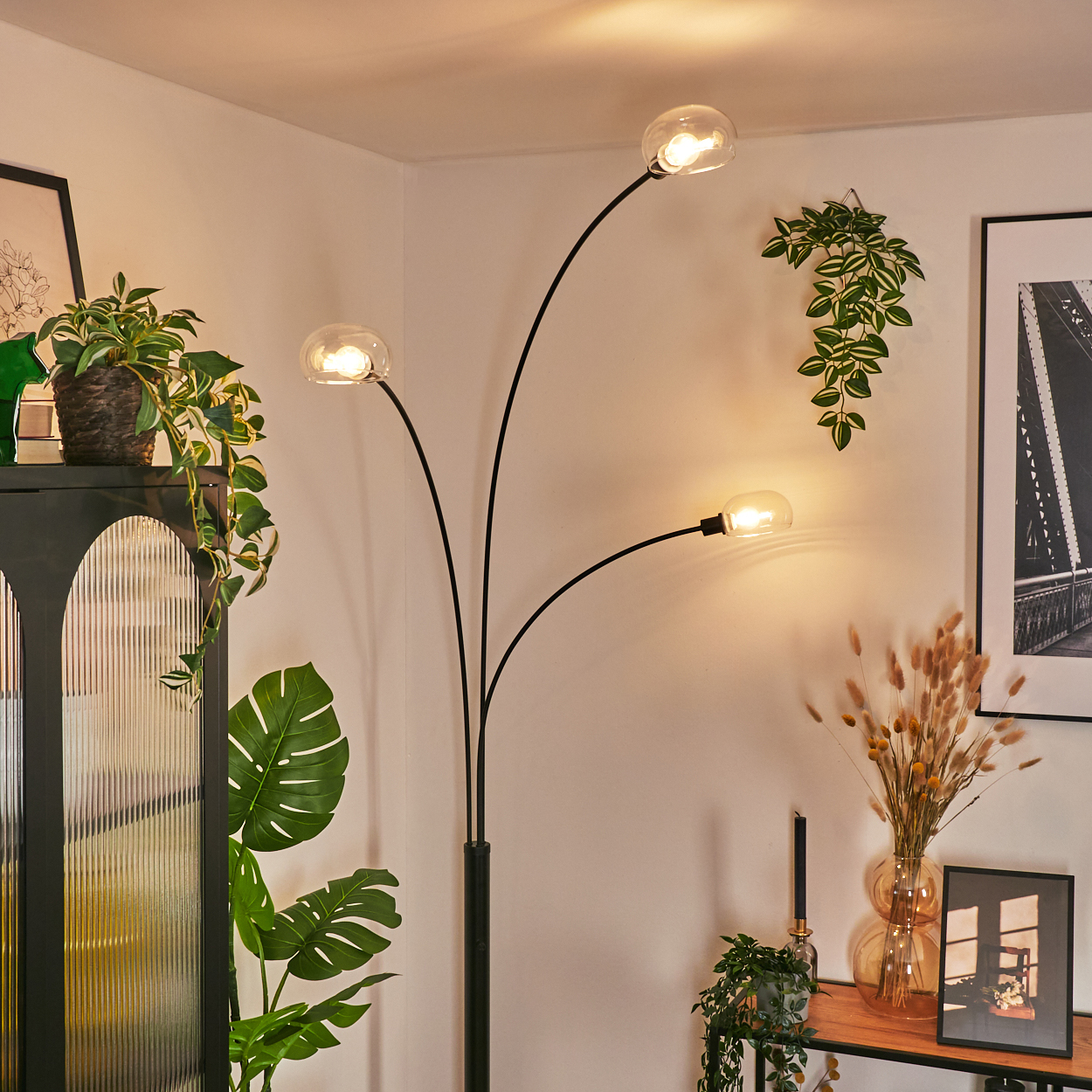 Ein modernes Wohnzimmer mit einer schwarzen Lampe mit drei Lichtern, Pflanzen und gerahmten Bildern. Warme Beleuchtung schafft eine gemütliche Atmosphäre.