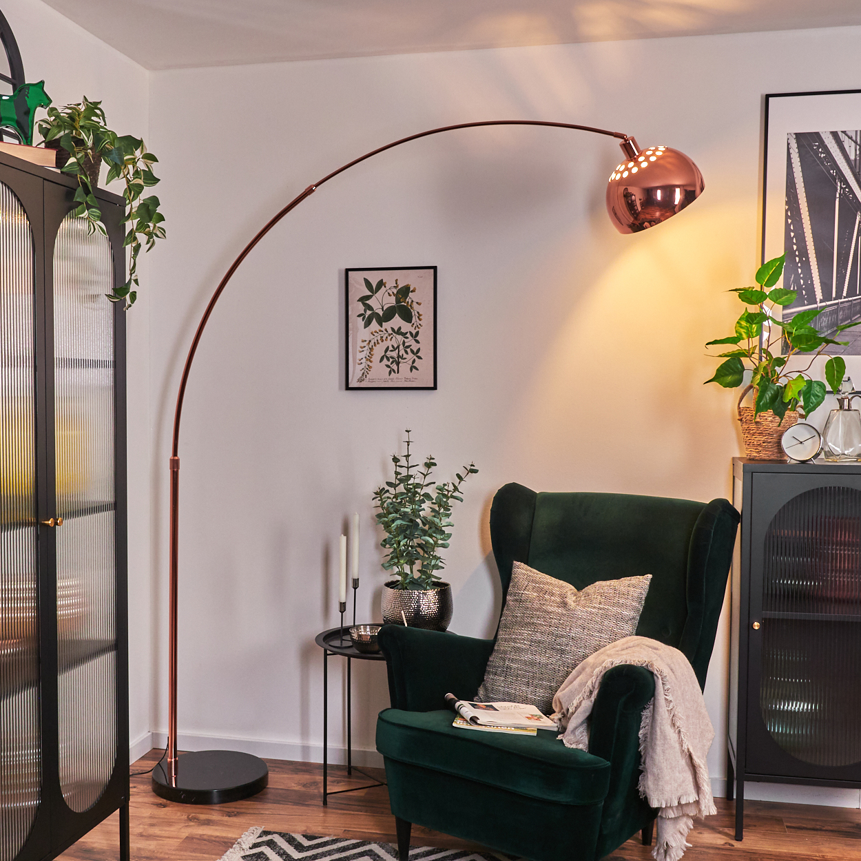 Ein Raum mit grünem Sessel, kupferfarbener Bogenlampe, schwarzem Schrank und Pflanzen. Ein Gemälde hängt an der Wand.