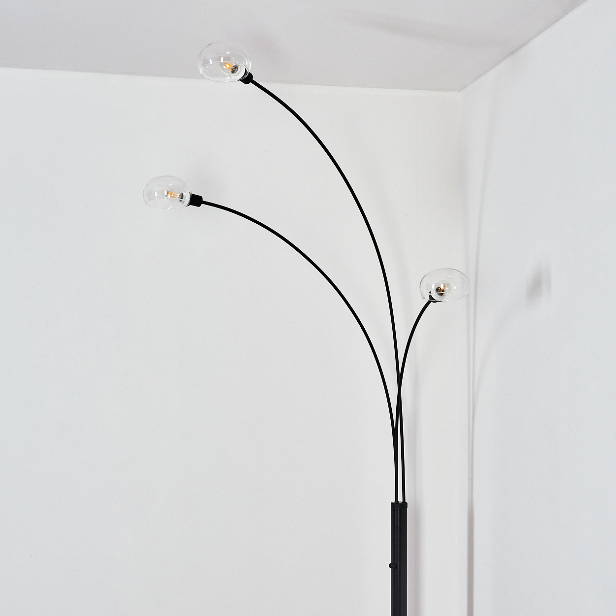 Eine moderne schwarze Stehlampe mit drei gebogenen Armen, die jeweils eine klare Glaskugelleuchte vor einer weißen Wand halten.