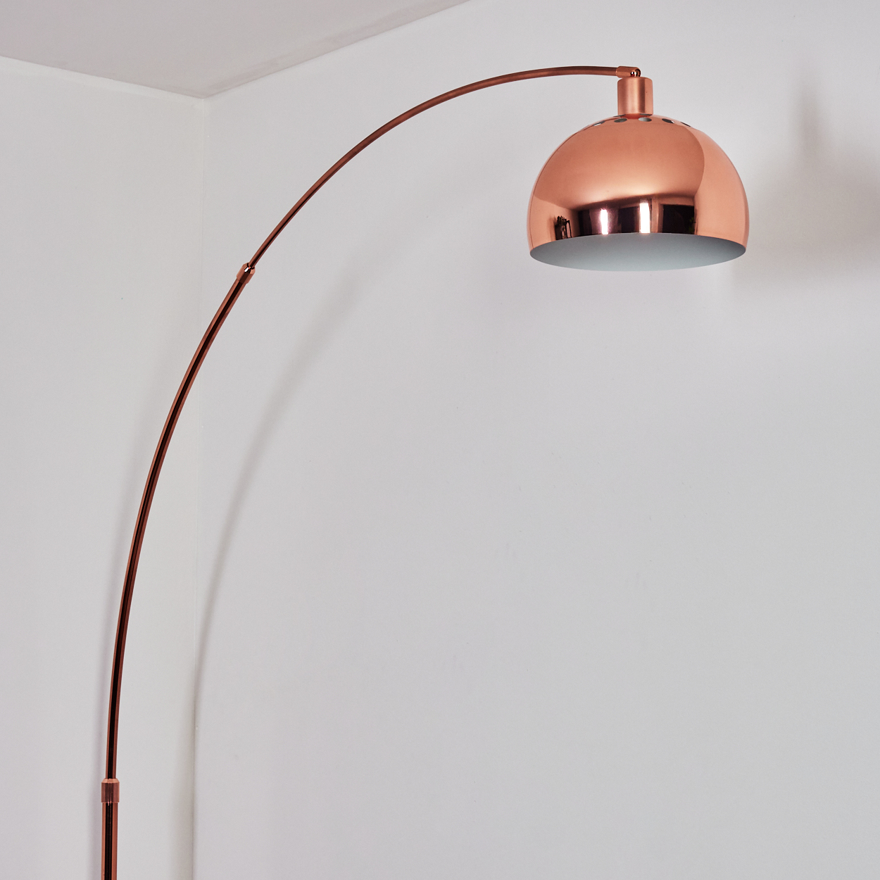 Eine kupferfarbene Bogenlampe steht vor einer weißen Wand. Die Lampe hat einen gebogenen Arm und einen kuppelförmigen Schirm.