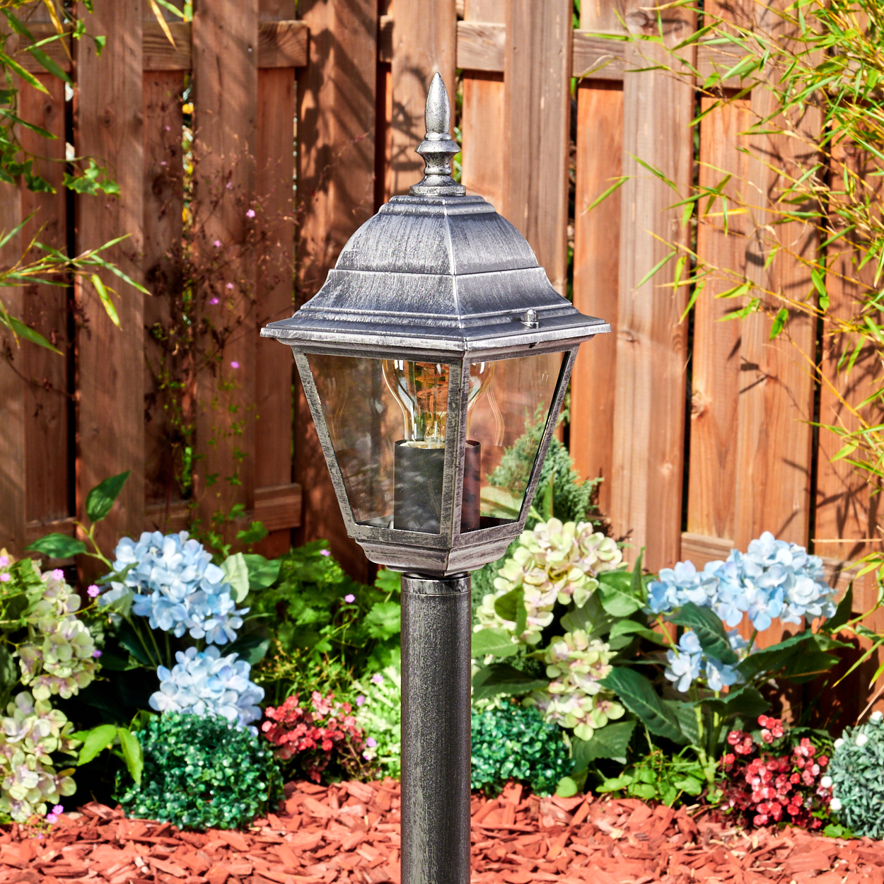 Eine dekorative Außenlampe steht in einem Garten mit blauen Hortensien und einem Holzzaun im Hintergrund.