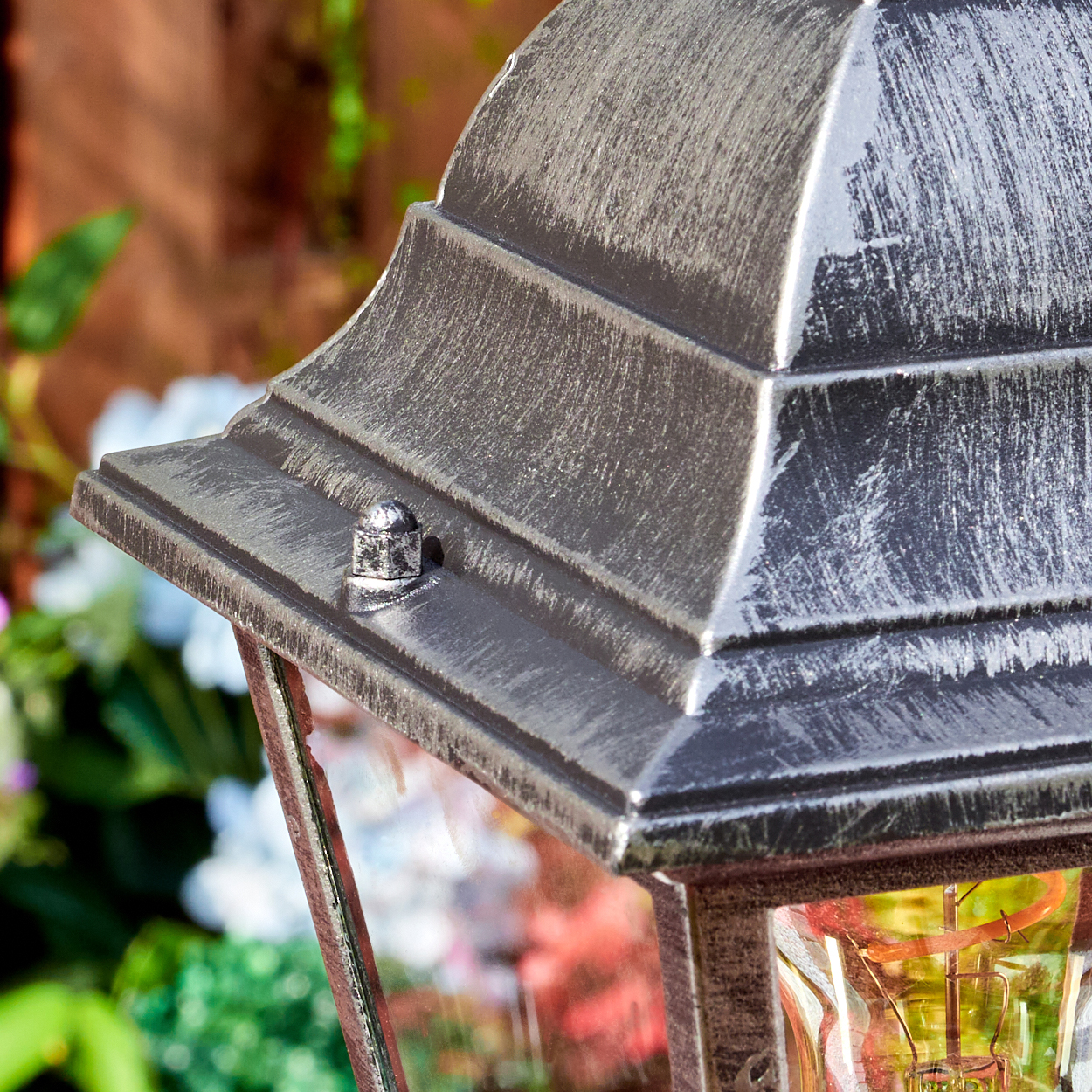 Nahaufnahme einer dekorativen Lampe mit Metallic-Finish. Die Lampe hat ein dachartiges Oberteil mit einem kleinen Knopf. Im Hintergrund sind Blumen zu sehen.