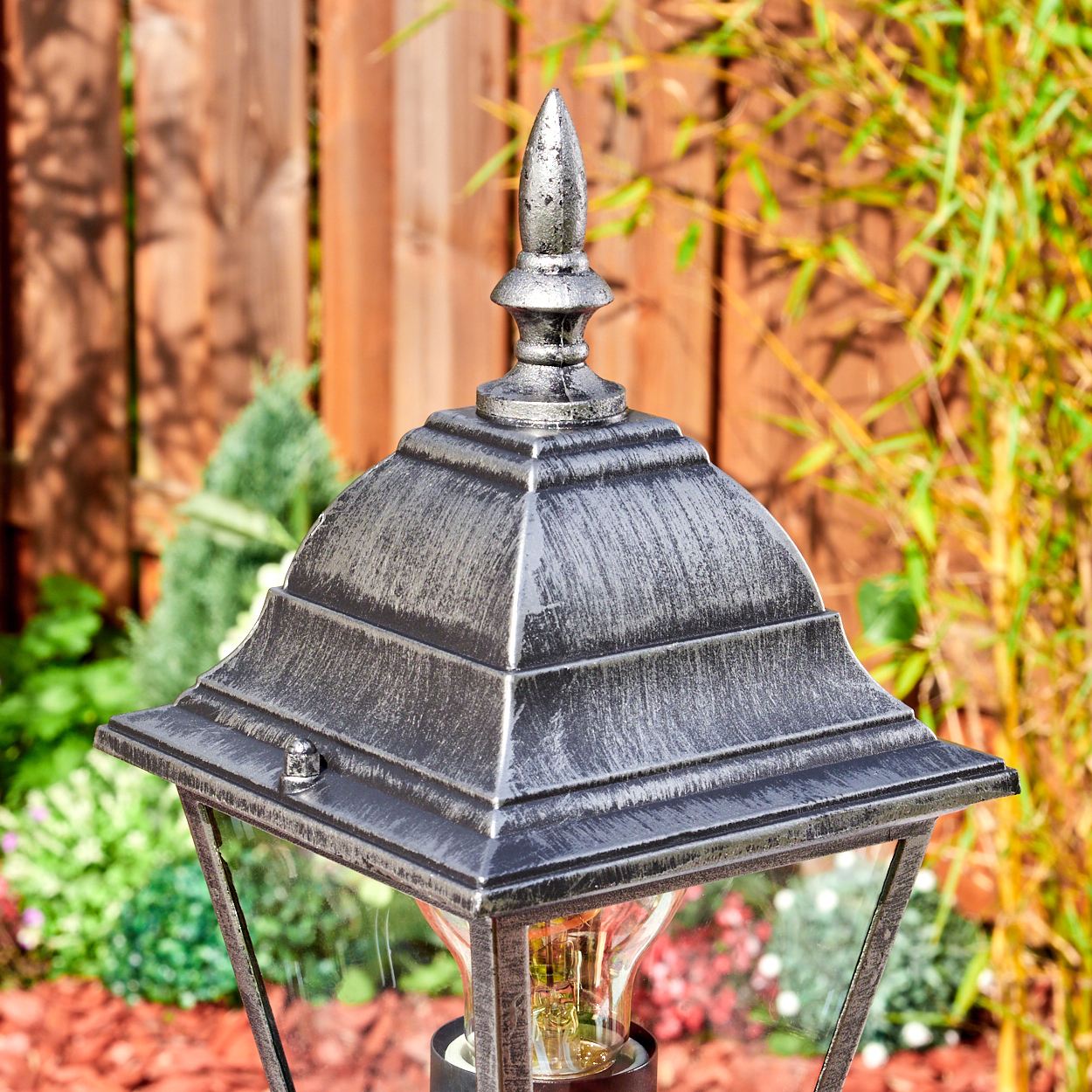 Eine dekorative Außenlampe mit silberner Oberseite. Dahinter ein Holzzaun und grüne Pflanzen. Die Lampe ist beleuchtet.