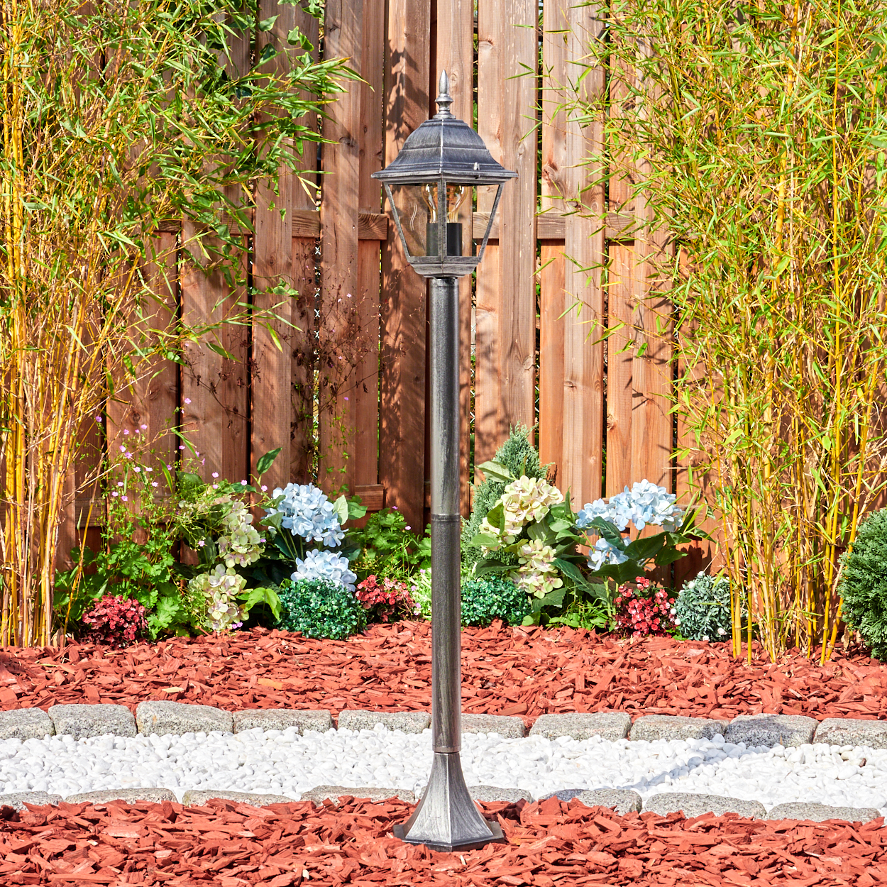 Eine dekorative Gartenlampe steht vor einem Holzzaun. Bambus und Blumen umgeben die Lampe. Rindenmulch und weiße Steine befinden sich im Vordergrund.