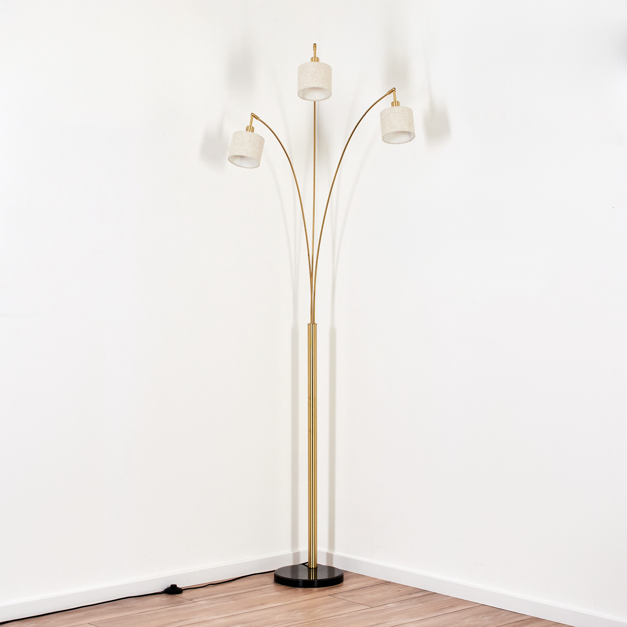 Eine goldene Stehlampe mit drei Lichtern. Sie steht auf einem schwarzen Sockel. Der Hintergrund ist eine weiße Wand und ein Holzboden.
