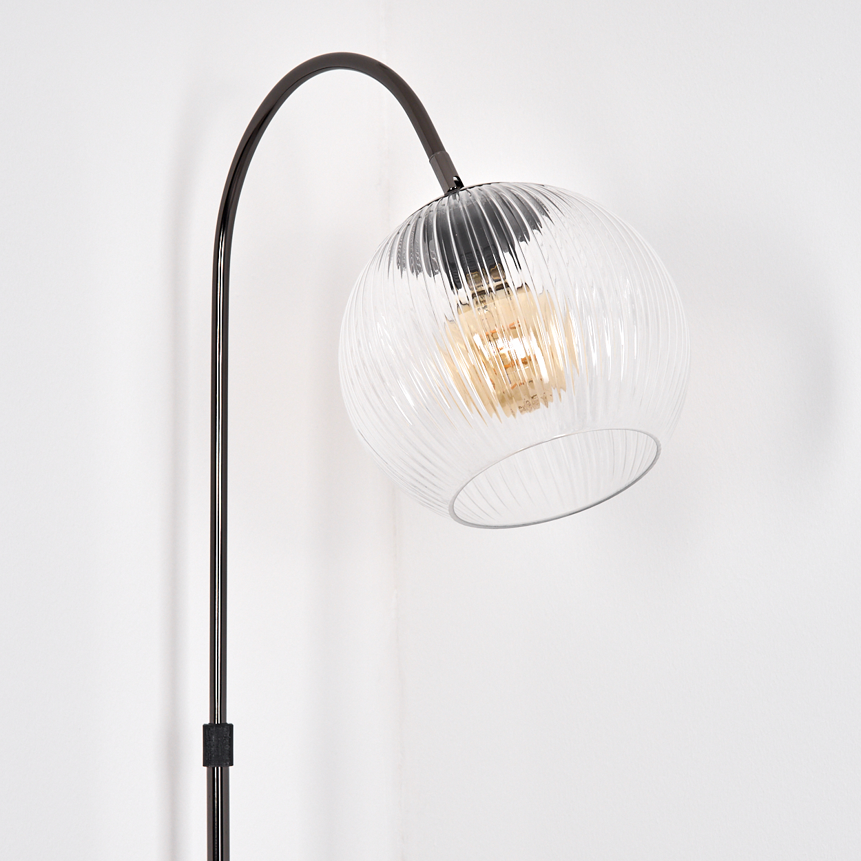Eine moderne Stehlampe mit einem durchsichtigen, gerippten Glaskugelschirm. Die Lampe hat einen gebogenen, dunklen Metallarm und eine gerade Stange. Weiße Wand.