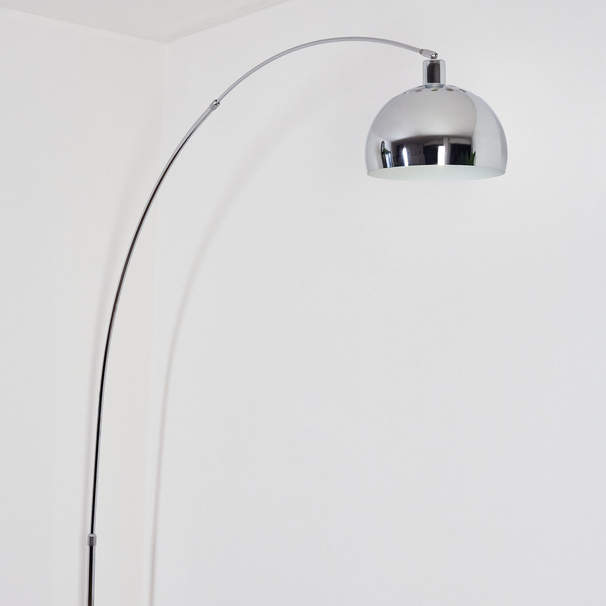 Eine moderne, verchromte Bogen-Stehlampe. Sie hat einen runden Lampenschirm und steht vor einer weißen Wand.