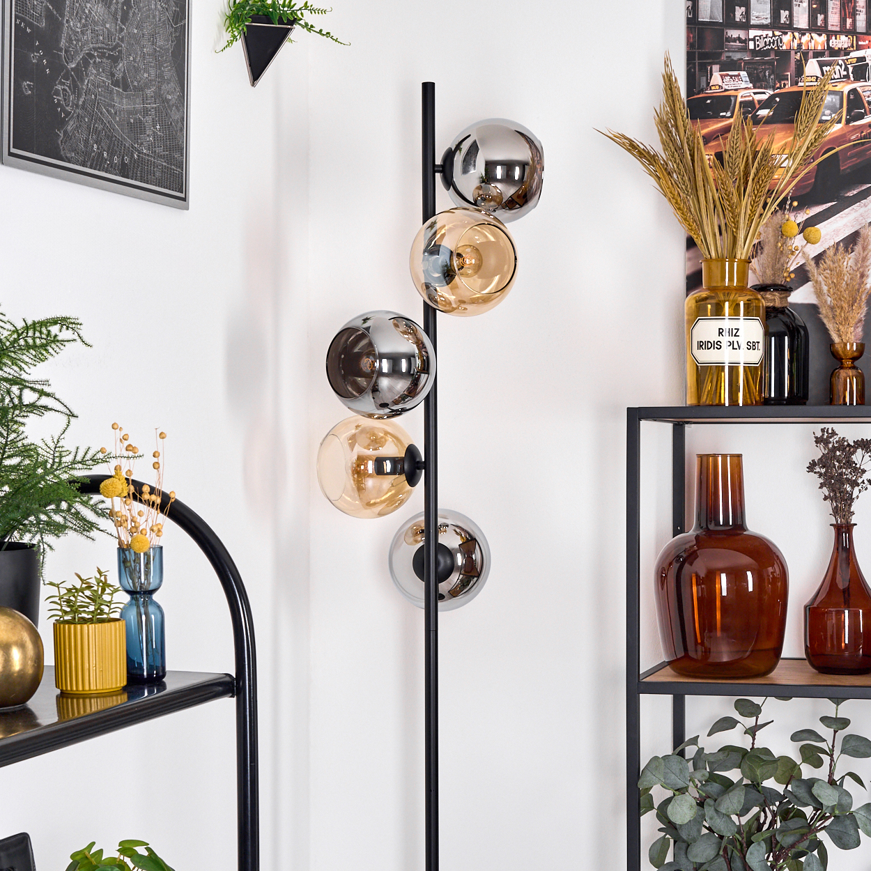 Ein modernes Interieur mit einer schwarzen Lampe, Glaskugeln, Regalen mit Pflanzen und Vasen sowie einer Karte und einem Taxibild an der Wand.