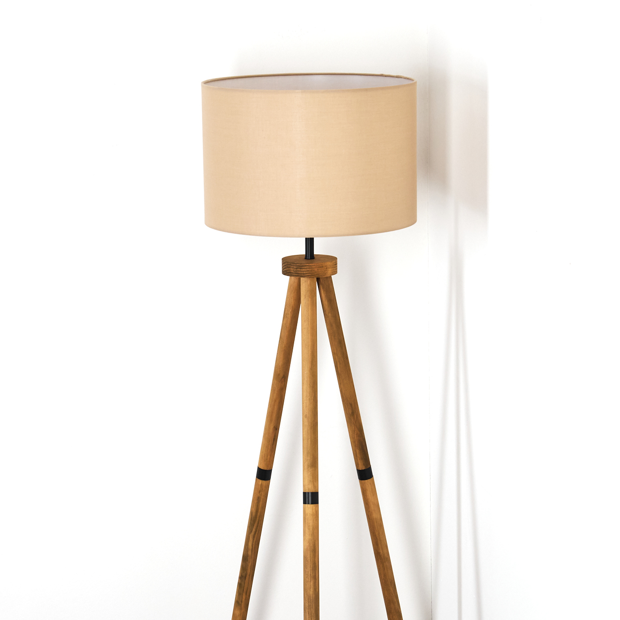 Ein beigefarbener Lampenschirm steht auf Holzstativbeinen vor einer weißen Wand. Die Lampe hat einen schwarzen Anschluss.