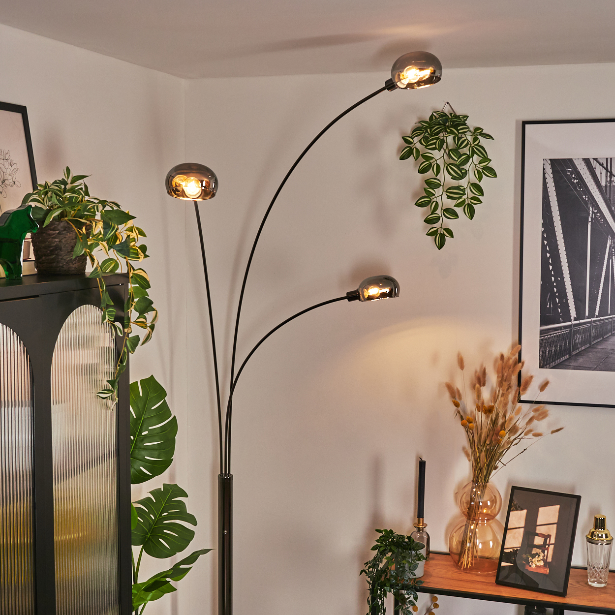 Ein modernes Wohnzimmer mit einer schwarzen Stehlampe mit drei Lichtern. Es gibt einen schwarzen Schrank, Pflanzen und ein Brückenbild.