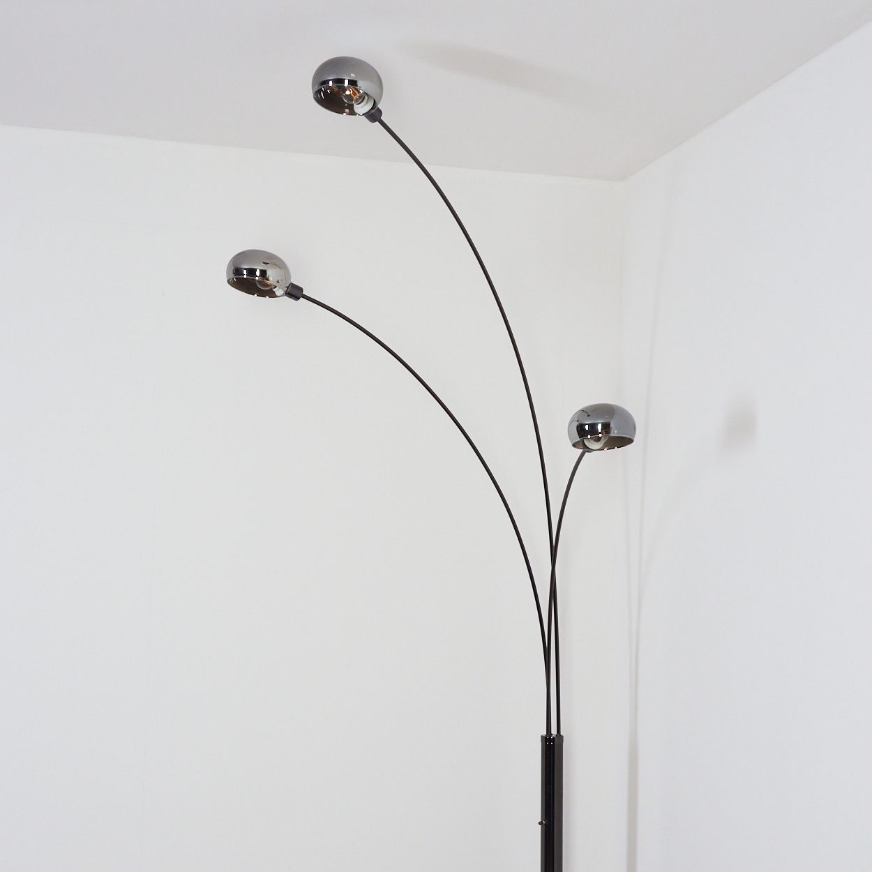 Eine moderne Stehlampe mit drei verchromten Kugellampen. Die Lampe hat schwarze, gebogene Stiele und steht in einer weißen Ecke.