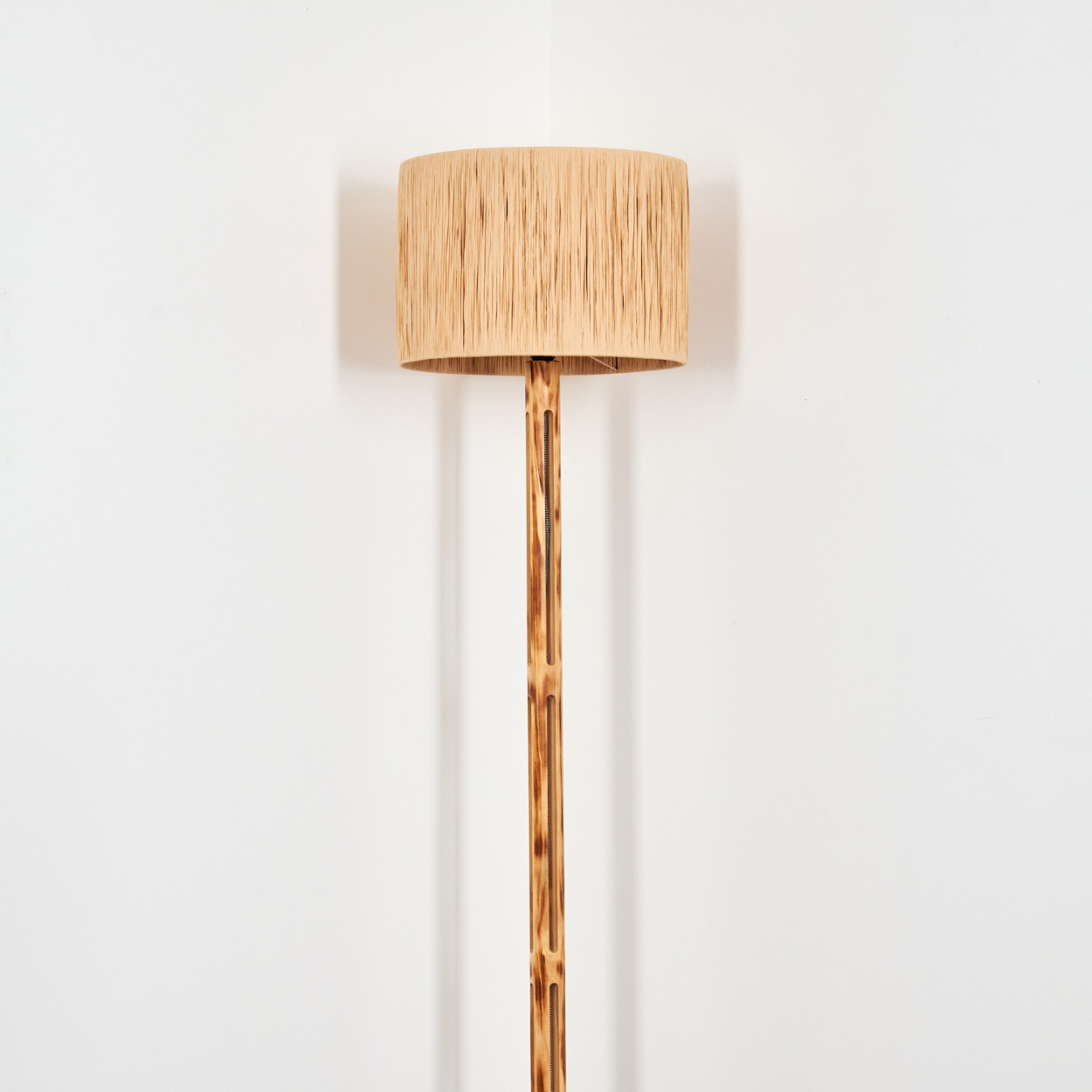 Eine Stehlampe steht in einer weißen Ecke. Sie hat einen beigefarbenen Lampenschirm und einen braunen Holzpfahl.