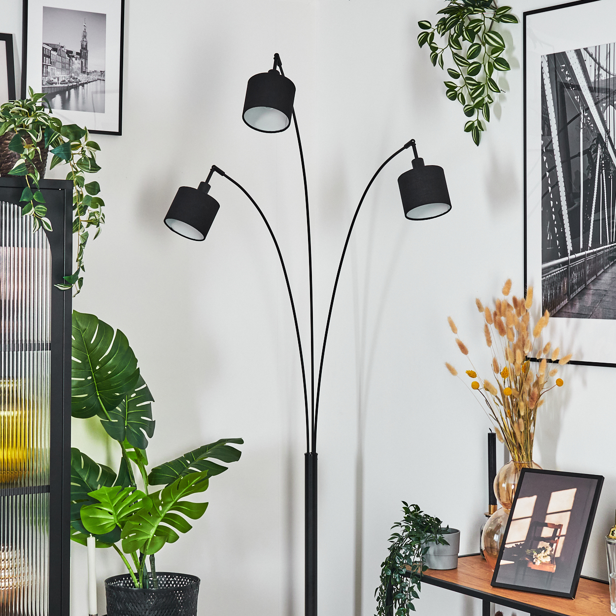 Eine moderne Raumszene mit einer schwarzen Stehlampe, Pflanzen, Bildern und einem Holzregal. Die Lampe hat drei Schirme. Pflanzen stehen in Töpfen.