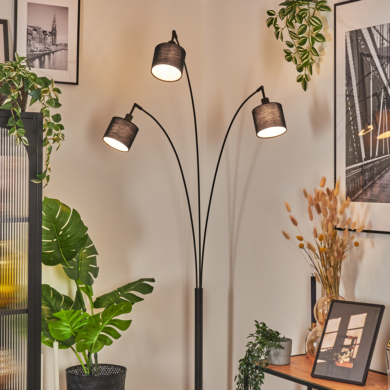 Eine schwarze Stehlampe mit drei Lichtern steht in einer Ecke. Pflanzen und Bilder schmücken den Raum. Ein Tisch ist sichtbar.