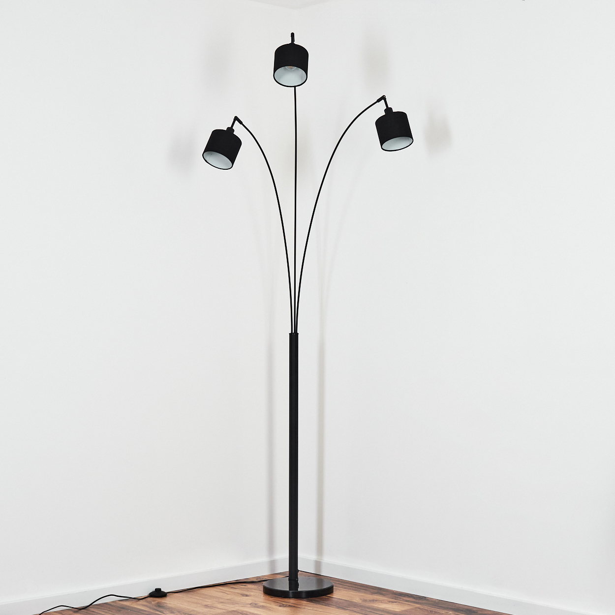 Eine schwarze Stehlampe mit drei Lampenschirmen. Sie steht in einer Ecke mit weißen Wänden und Holzboden.