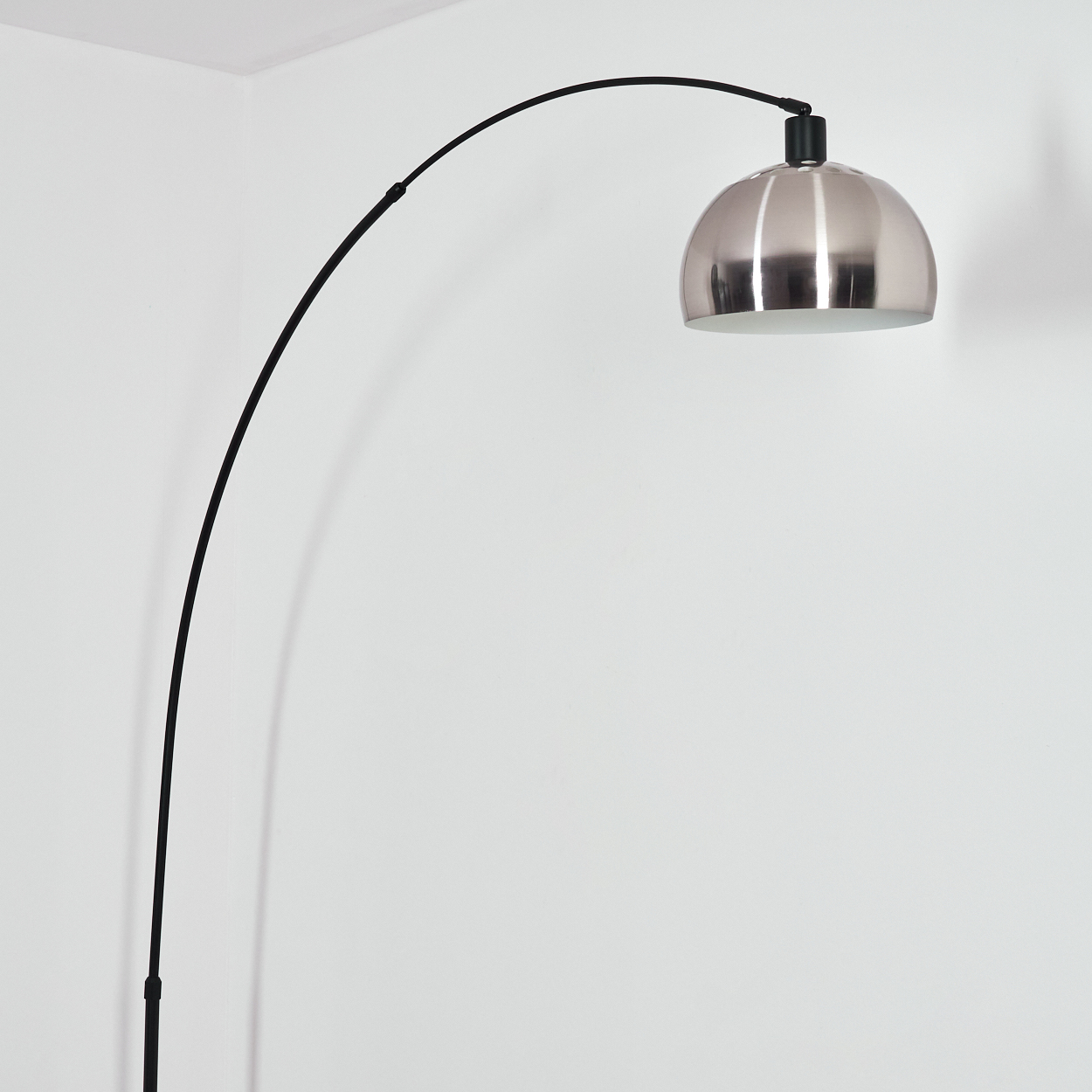 Eine moderne Bogenstehlampe mit silbernem Kuppelschirm. Der schwarze Arm verläuft über einer weißen Wand.