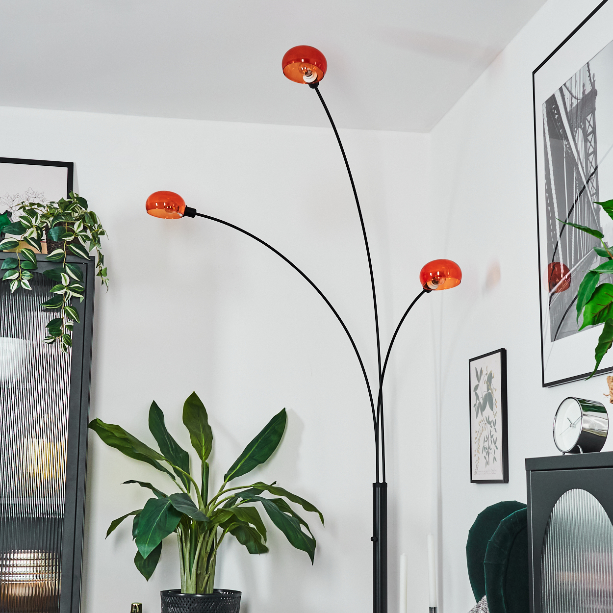 Ein modernes Wohnzimmer mit einer schwarzen Stehlampe mit drei orangefarbenen Kugellampen. Es gibt Pflanzen, Bilder und eine Uhr.