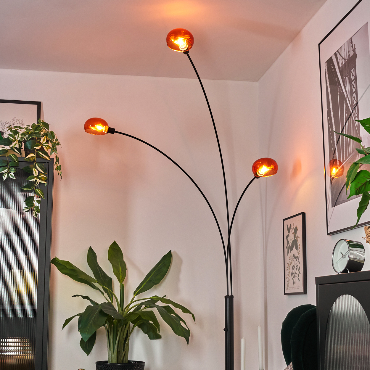 Ein modernes Wohnzimmer mit einer schwarzen Stehlampe mit orangefarbenen Glühbirnen, Pflanzen und gerahmter Kunst. Der Raum ist warm beleuchtet.