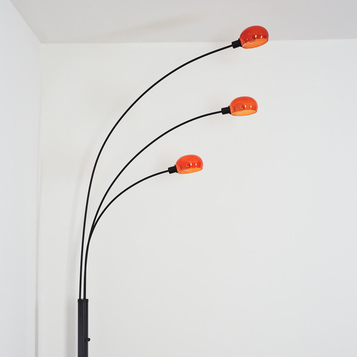 Eine moderne Stehlampe mit drei gebogenen schwarzen Armen, die jeweils eine orange-rote Kugellampe vor einer weißen Wand halten.