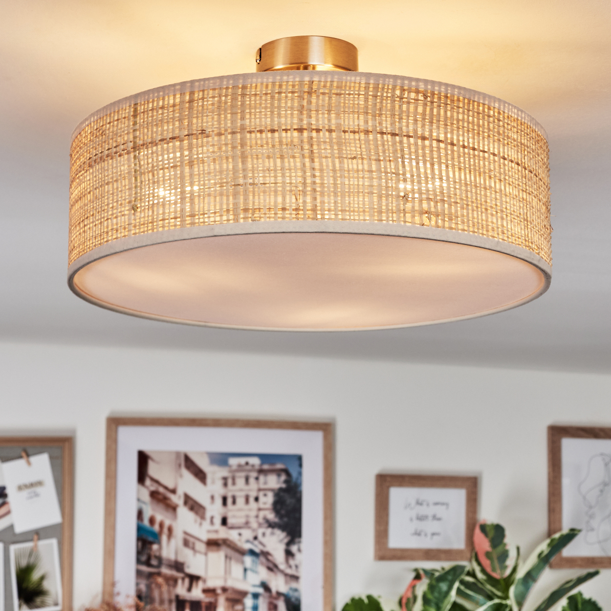 Ein gewebter Lampenschirm hängt von der Decke. Darunter sind gerahmte Bilder und eine Pflanze zu sehen. Das Licht ist warm.