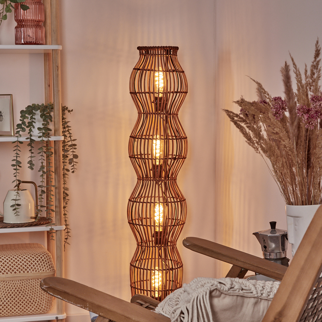 Eine gewebte Stehlampe aus Rattan ist beleuchtet, mit einem Holzstuhl und Pflanzen im Hintergrund. Eine Kaffeekanne ist ebenfalls sichtbar.