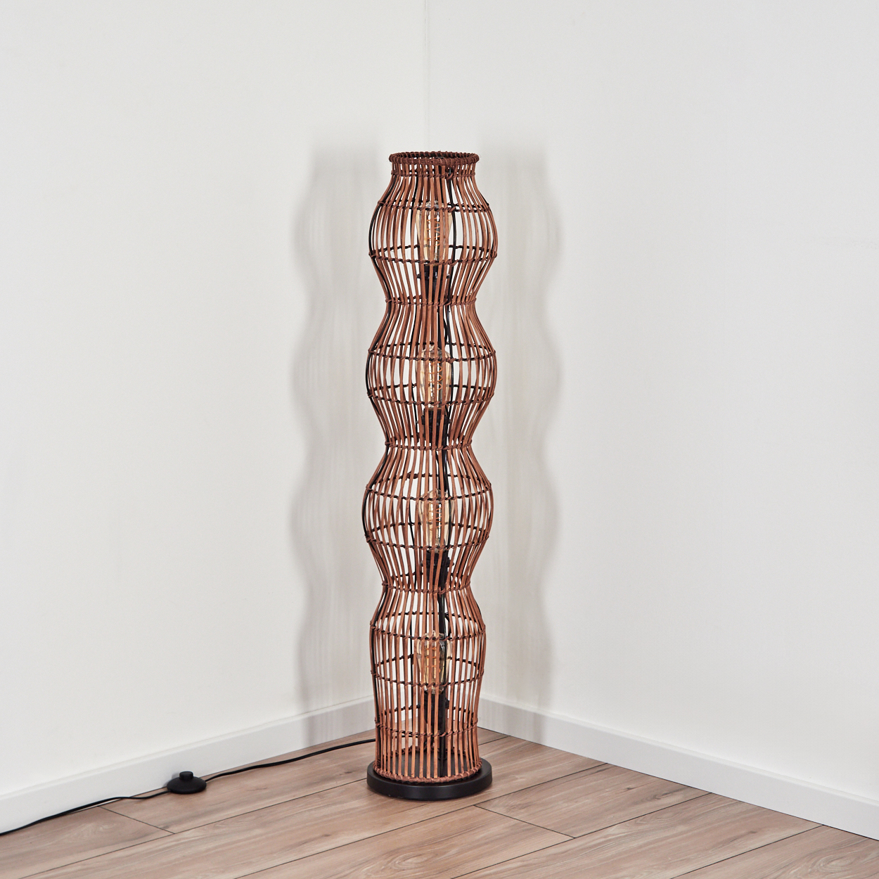 Eine hohe, braune gewebte Lampe steht in einer Ecke. Die Lampe hat eine Glühbirne. Sie steht auf einem Holzboden mit weißen Wänden.