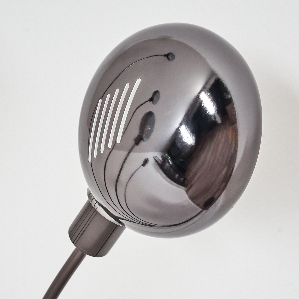 Nahaufnahme eines metallischen Lampenkopfes mit weißen Lüftungsschlitzen, der einen Raum reflektiert. Die Lampe steht auf einem dunklen Stiel vor weißem Hintergrund.