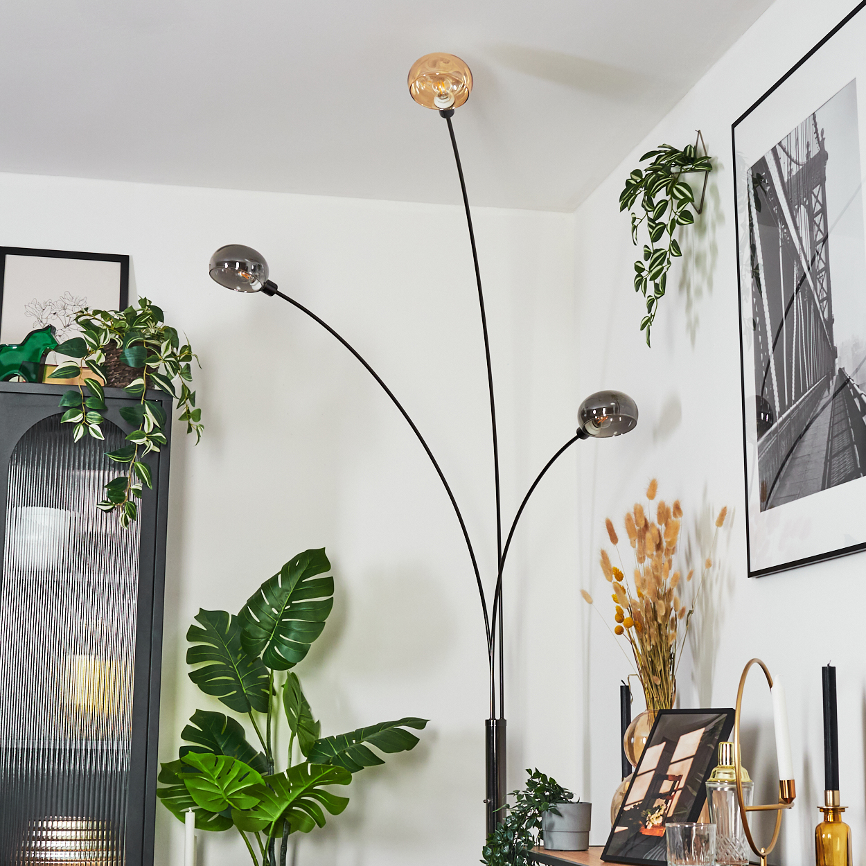 Eine moderne Stehlampe mit drei Glaskugelleuchten. Pflanzen, Bilder und Dekorationsartikel sind um die Lampe angeordnet.
