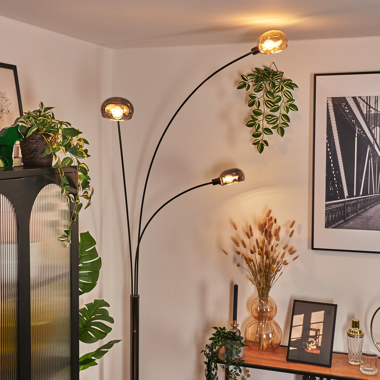 Ein modernes Wohnzimmer mit schwarzer Stehlampe, Pflanzen, schwarzem Schrank und gerahmtem Brückenfoto. Warme Beleuchtung schafft eine gemütliche Atmosphäre.
