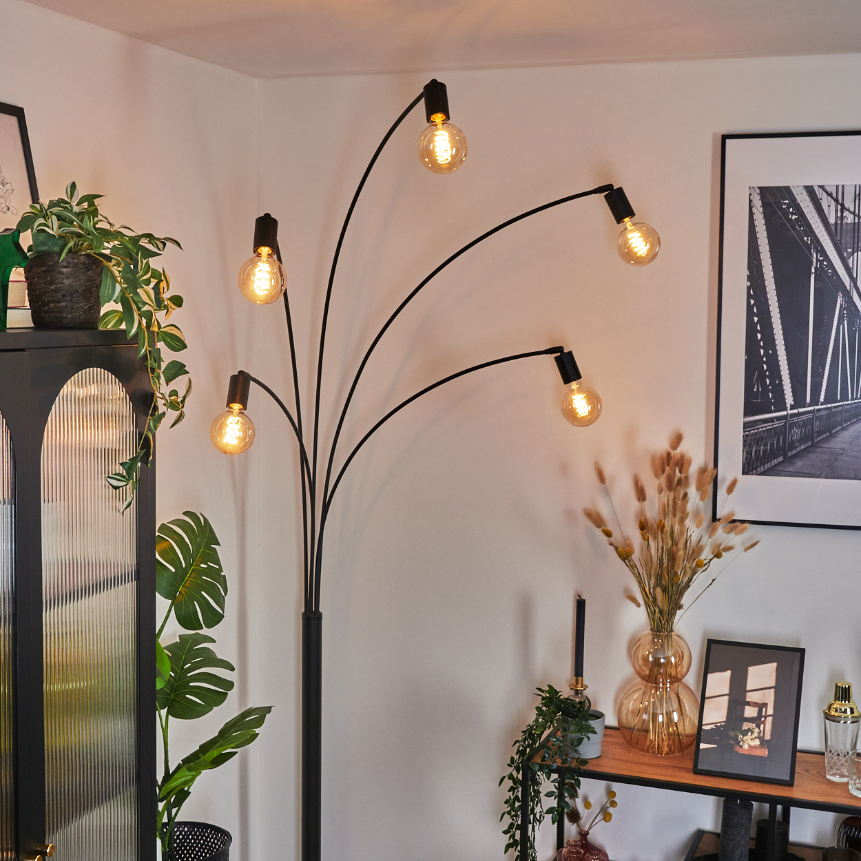 Eine moderne Stehlampe mit fünf Glühbirnen. Pflanzen und ein gerahmtes Bild schmücken den Raum, mit einem schwarzen Schrank und einem Holztisch.