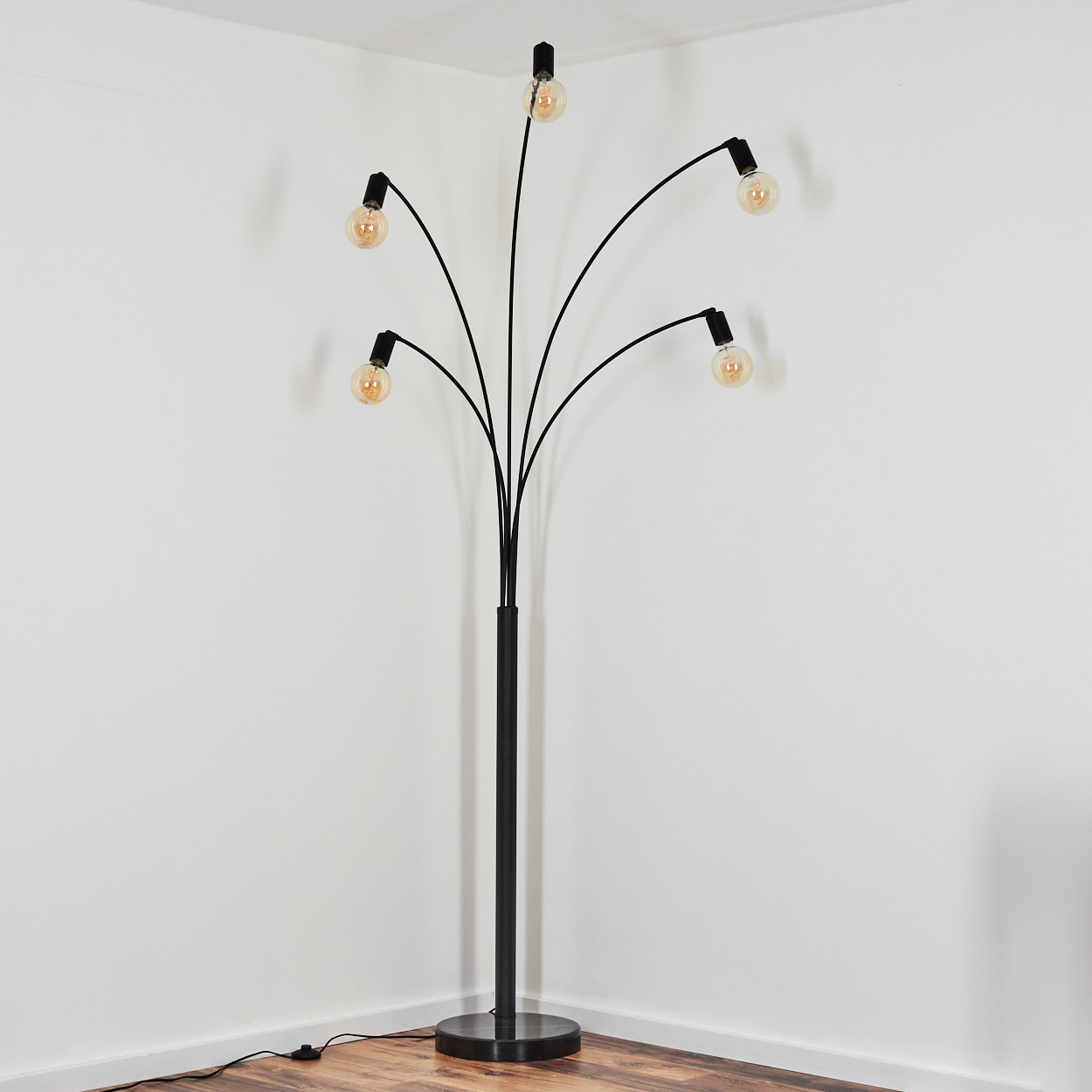Eine schwarze Stehlampe mit sechs Glühbirnen, in einem Bogen angeordnet, steht in einer Ecke. Der Hintergrund ist eine weiße Wand und ein Holzboden.