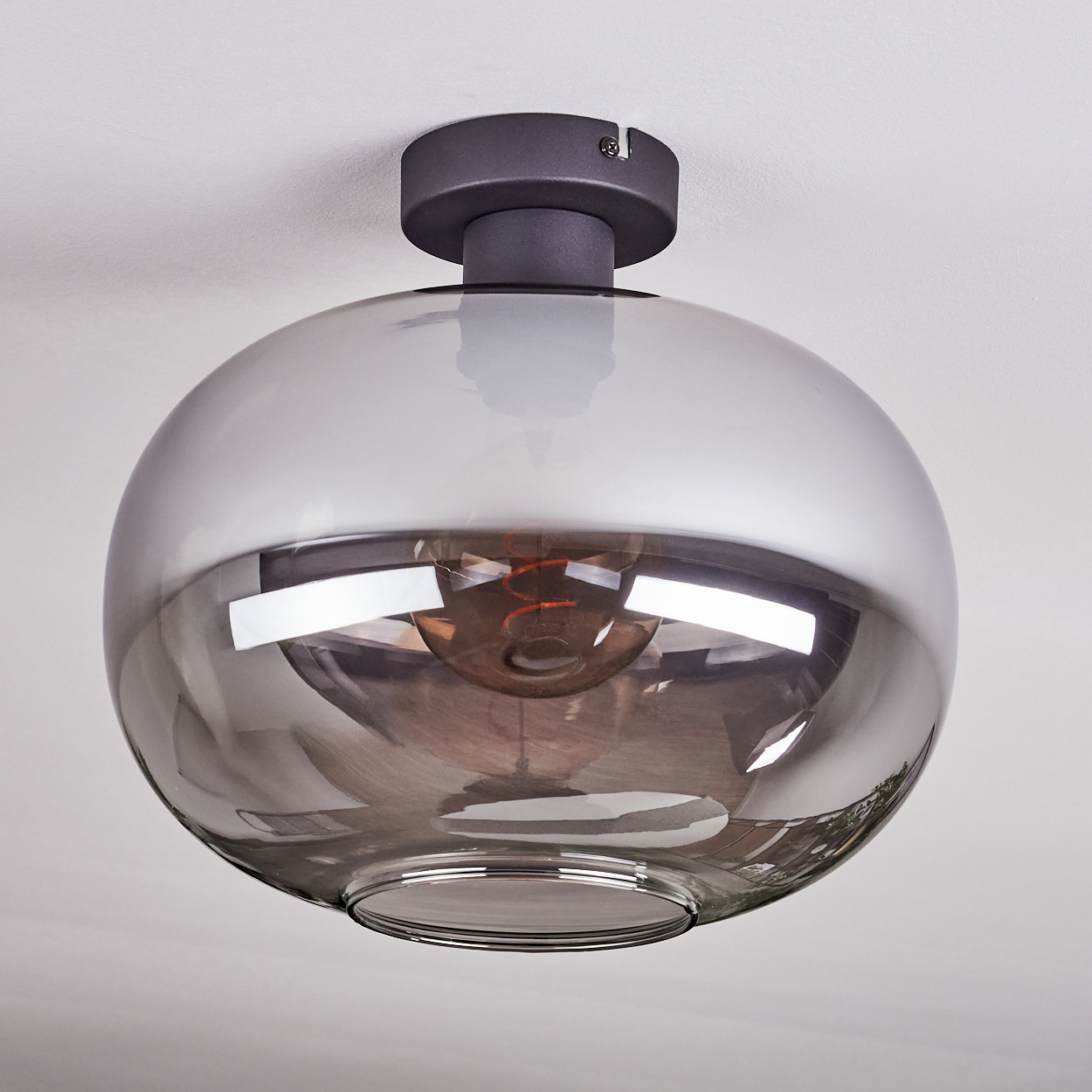 Moderne Deckenlampe aus grau getöntem Glas mit dunklem Metallsockel, an einer weißen Decke hängend, mit sichtbarer Glühbirne.
