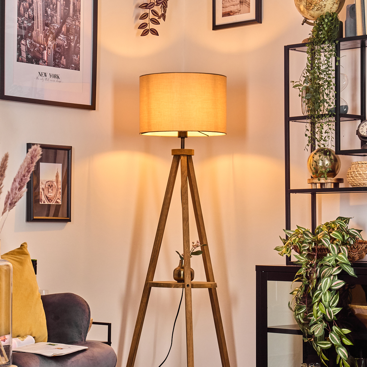 Ein Raum mit einer hölzernen Stativlampe, gerahmten Bildern und einem schwarzen Regal mit Pflanzen. Ein Sofa und Dekorationsartikel sind ebenfalls zu sehen.